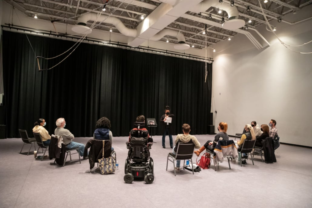 A small group attends an accessible theatre program in a black-box performance space, with audience members seated in a circle and one participant using a power wheelchair.