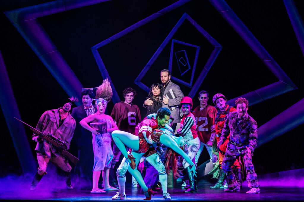 A brightly costumed ensemble reacts around two performers striking a dramatic dance pose at center stage, framed by purple lighting, fog and geometric set pieces.
