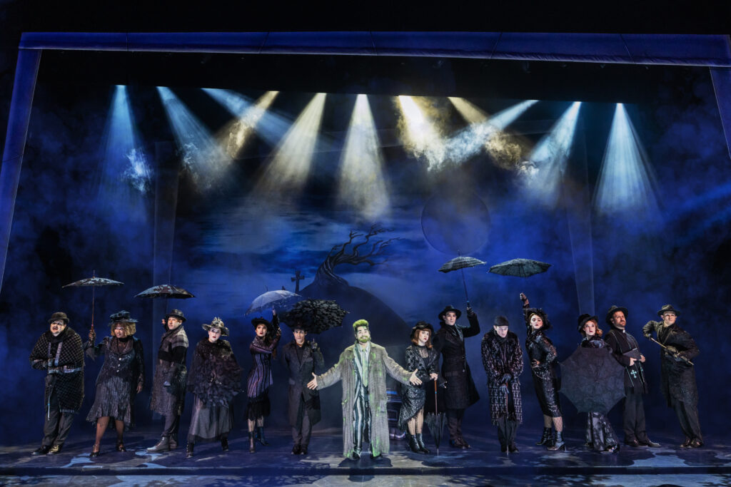 A large ensemble dressed in dark Victorian mourning clothes stands beneath dramatic blue and white spotlights, surrounding a green-haired performer in a striped suit.
