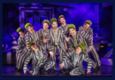 Nine performers in matching black-and-white striped suits and green-streaked hair pose energetically on a purple-lit stage filled with theatrical fog.