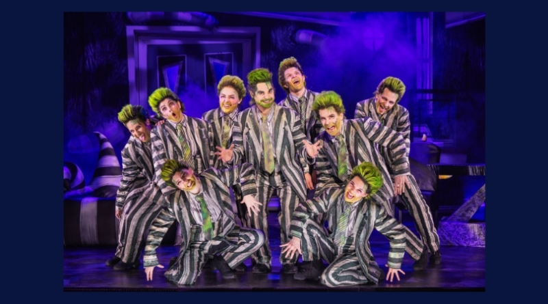 Nine performers in matching black-and-white striped suits and green-streaked hair pose energetically on a purple-lit stage filled with theatrical fog.