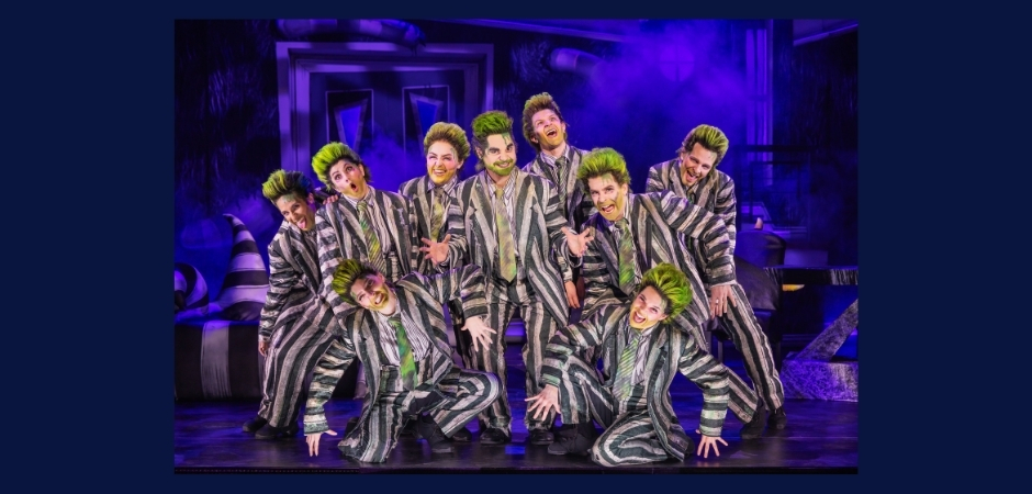 Nine performers in matching black-and-white striped suits and green-streaked hair pose energetically on a purple-lit stage filled with theatrical fog.