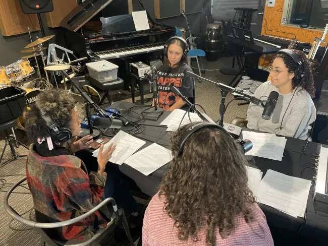 Four people wearing headphones sit around a table with microphones and notes during a podcast recording in a music studio filled with instruments.
