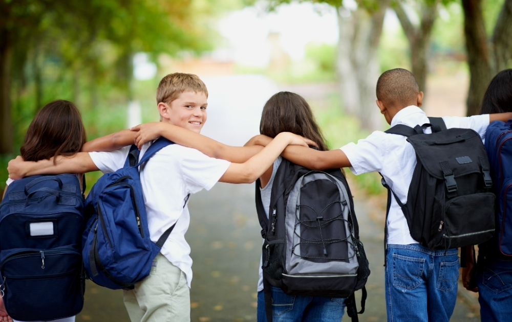 A diverse group of school-age children wearing backpacks walk together outdoors with their arms around one another.