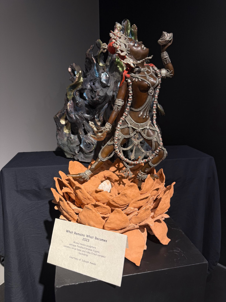 Detailed mixed-media sculpture featuring an ornate bronze figure standing on a layered orange ceramic base, displayed against a dark backdrop in the Heritage in Motion exhibition.