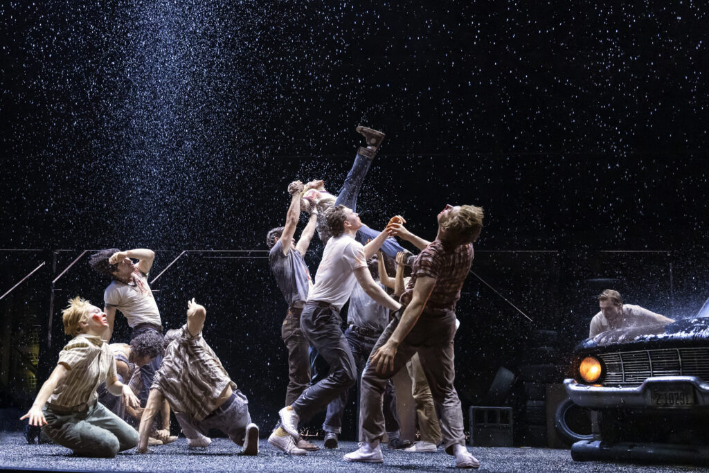 Cast members perform a highly physical fight sequence in the rain, with bodies lifted and thrown into motion as water falls across the darkened stage.