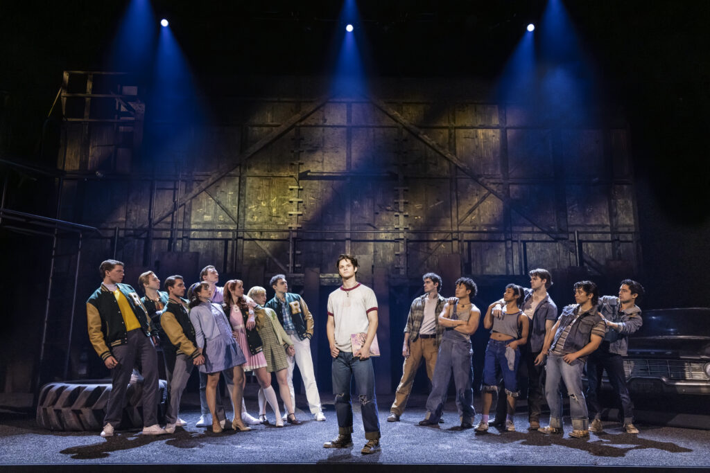 A young man stands center stage holding a book as cast members from “The Outsiders” gather on either side beneath blue spotlights, visually separating the Socs and Greasers against the production’s industrial Tulsa set.
