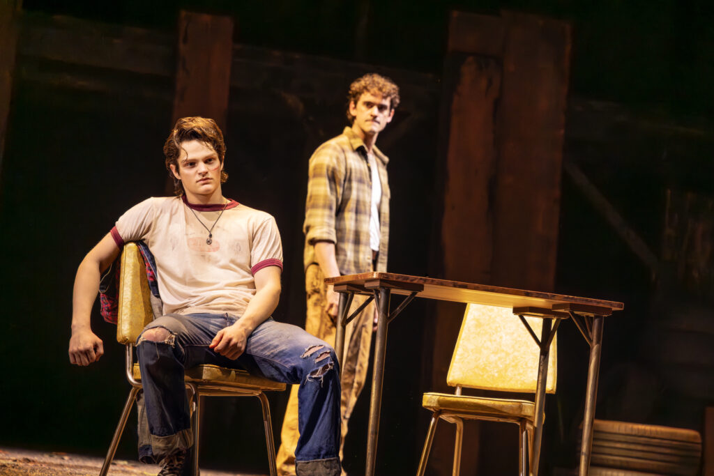 A young man with visible facial injuries sits in a chair in a tense scene from “The Outsiders,” while another young man stands behind him near a small table on the dimly lit stage.
