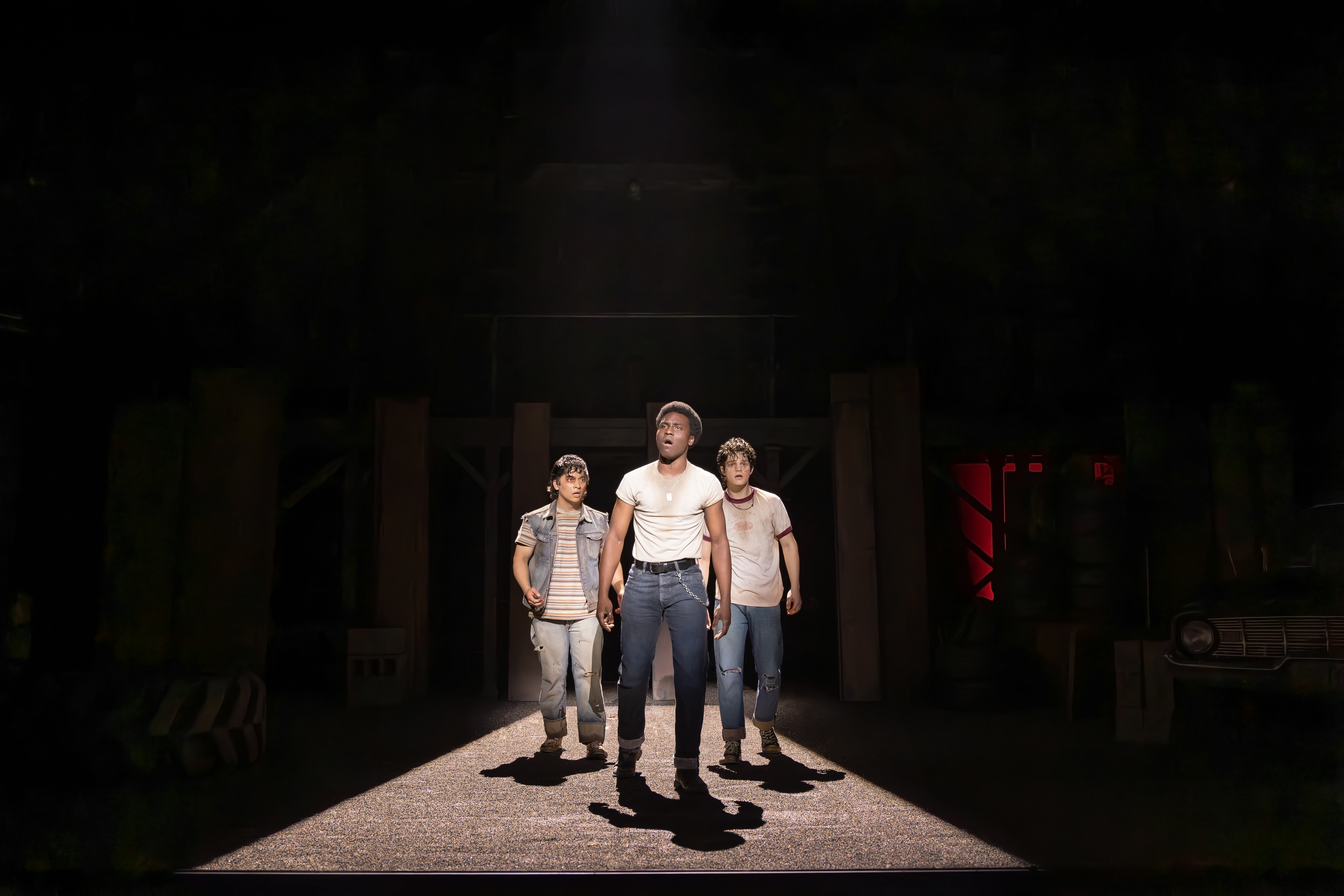 Three performers from “The Outsiders” stand in a narrow spotlight on a darkened stage, with one man positioned at center and two others just behind him as a vintage car and industrial set pieces fade into the shadows.