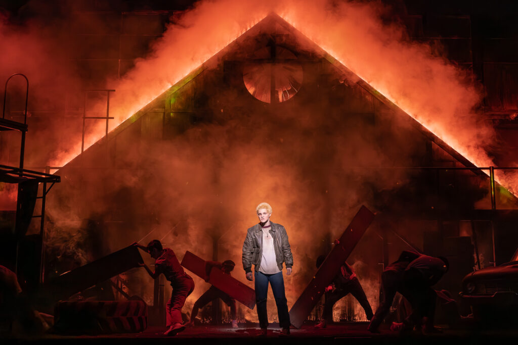 A performer stands center stage in “The Outsiders” as flames and heavy smoke engulf the set behind him, with other cast members moving through the fiery scene.