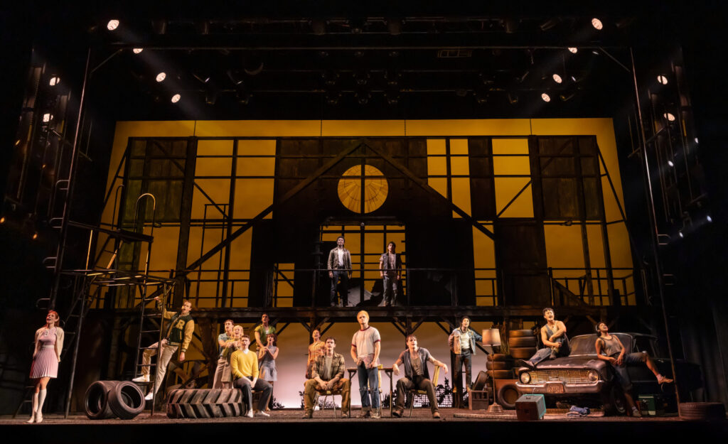 The full cast of “The Outsiders” gathers across a large industrial-style set framed by warm amber lighting, scaffolding, tires and a vintage car.