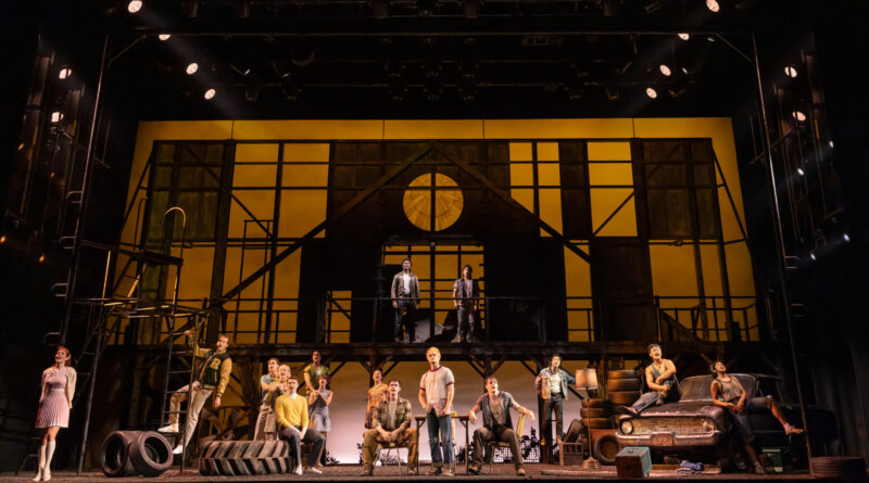 The full cast of “The Outsiders” gathers across a large industrial-style set framed by warm amber lighting, scaffolding, tires and a vintage car.