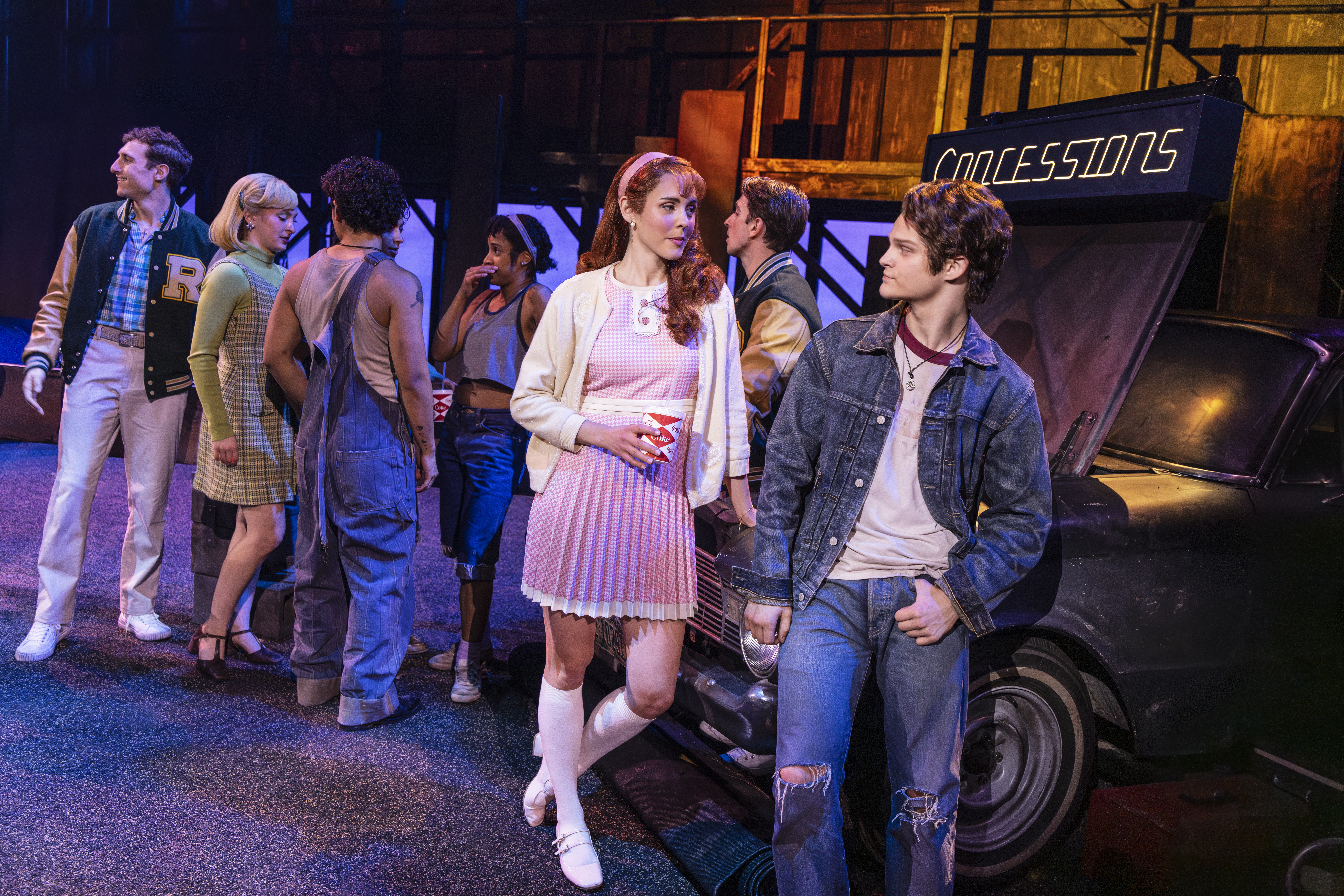 A young woman in a pink dress and a young man in a denim jacket face one another near a drive-in concession stand as other cast members mingle in the background.