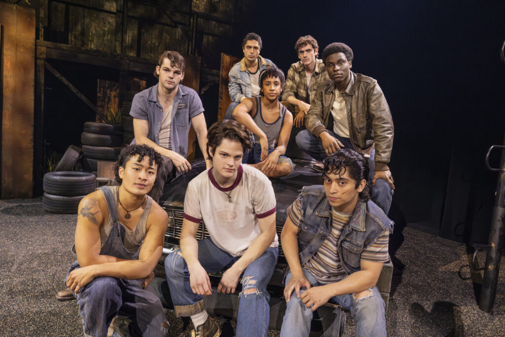 Members of the Greasers gather around and on top of a vintage car, posing together against the production’s industrial East Tulsa set.