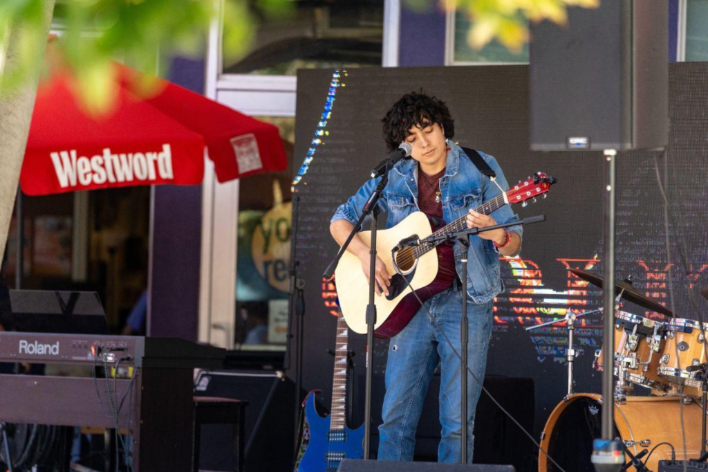 A musician in a denim jacket and jeans plays an acoustic guitar and sings into a microphone on an outdoor stage, with a keyboard, drum kit and additional guitars nearby. A red Westword umbrella is visible in the background.