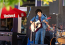 A musician in a denim jacket and jeans plays an acoustic guitar and sings into a microphone on an outdoor stage, with a keyboard, drum kit and additional guitars nearby. A red Westword umbrella is visible in the background.