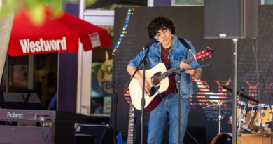 A musician in a denim jacket and jeans plays an acoustic guitar and sings into a microphone on an outdoor stage, with a keyboard, drum kit and additional guitars nearby. A red Westword umbrella is visible in the background.