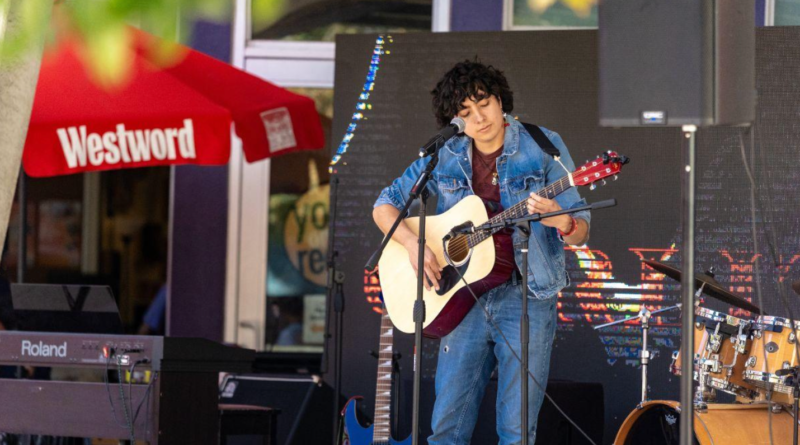 A musician in a denim jacket and jeans plays an acoustic guitar and sings into a microphone on an outdoor stage, with a keyboard, drum kit and additional guitars nearby. A red Westword umbrella is visible in the background.