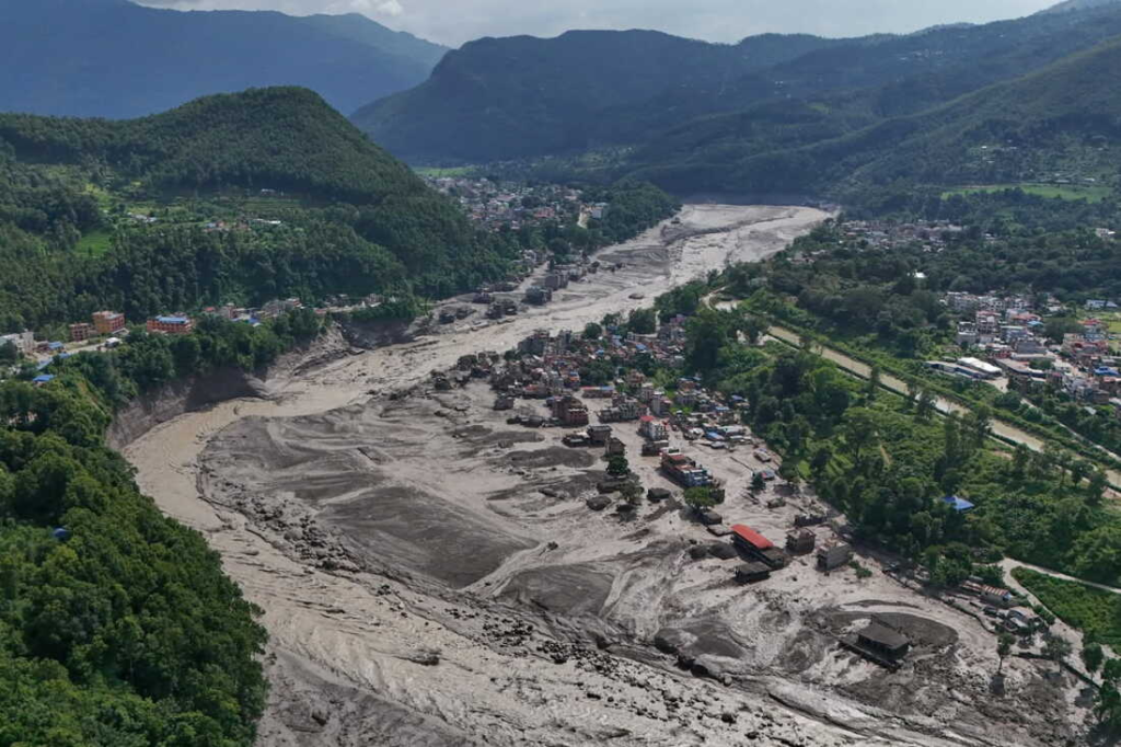 Aerial view of a mountainous community in Nepal showing extensive flood and debris damage, with mud and sediment covering a wide river corridor and surrounding homes and buildings.