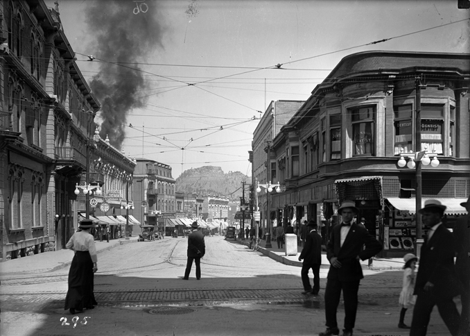 Black-and-white historical photograph of a Colorado downtown street lined with commercial buildings, pedestrians and streetcars, with smoke rising in the distance.