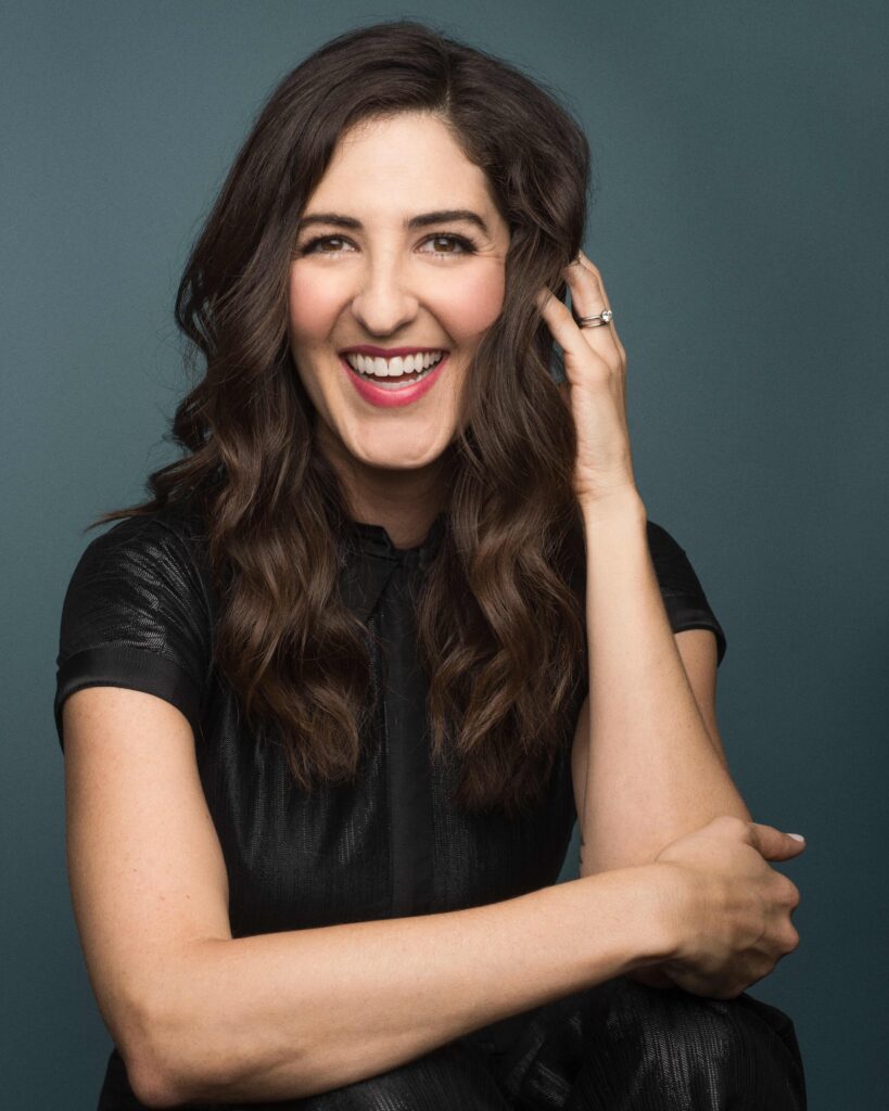 Portrait of actor D’Arcy Carden smiling in a black outfit, with one hand resting against her hair. Photo by Luke Fontana.