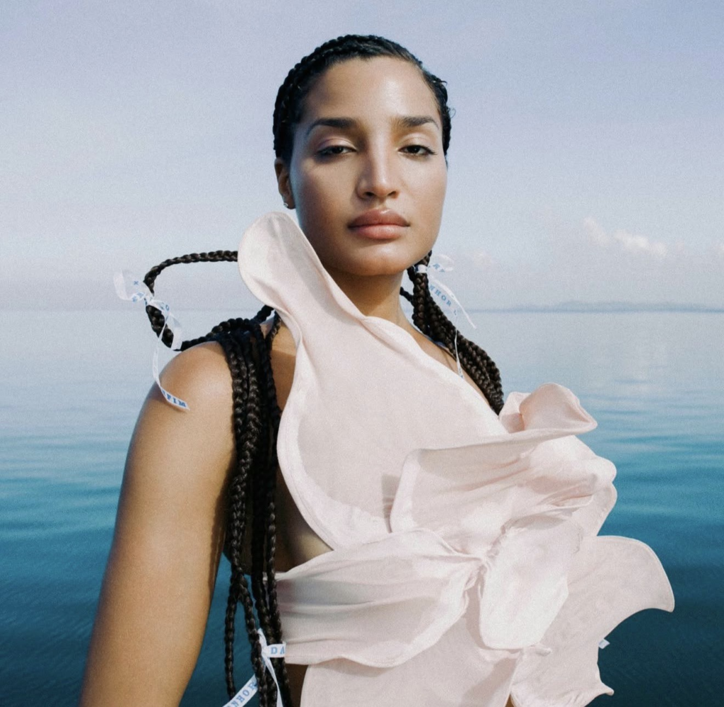 Portrait of actor and model Indya Moore wearing a pale pink sculptural dress with long braided hair, photographed against a blue ocean backdrop.