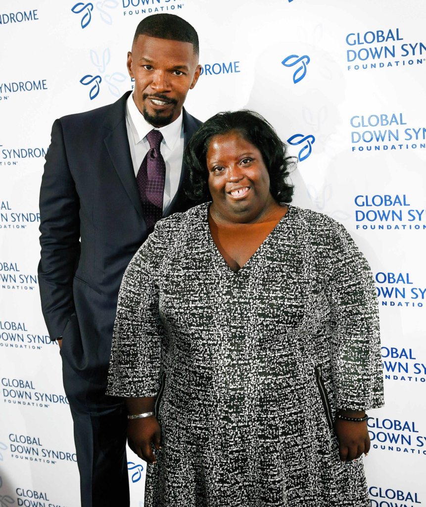 Actor Jamie Foxx stands beside his sister, DeOndra Dixon, smiling in front of a Global Down Syndrome Foundation backdrop.