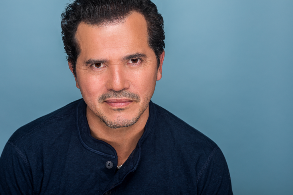 Headshot of actor John Leguizamo wearing a dark blue shirt against a light blue studio background. 