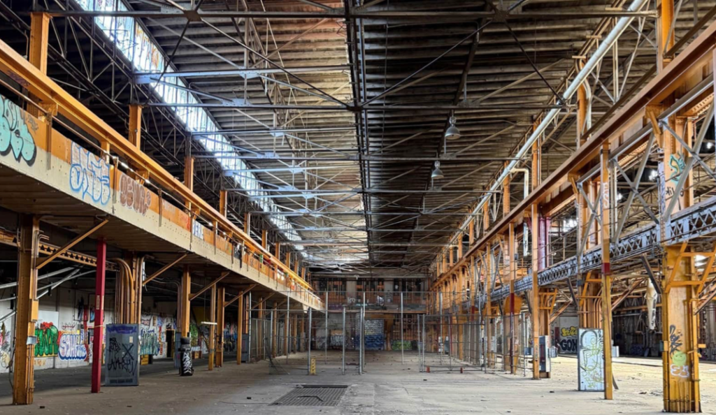 Interior of a former Burnham Yard industrial building in Denver, featuring exposed steel beams, overhead walkways, graffiti-covered walls and a large open factory floor.