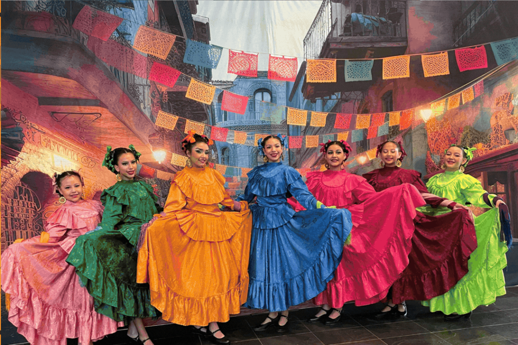 Seven dancers wearing vibrant, traditional Mexican folklórico dresses pose beneath colorful papel picado decorations against a festive streetscape backdrop.
