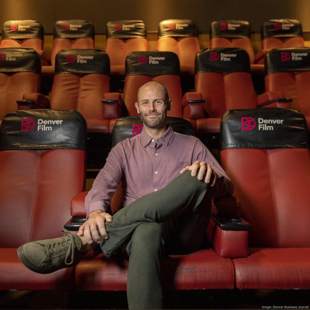 Denver Film CEO Kevin Smith sits cross-legged in a red theater seat, surrounded by rows of seats featuring the Denver Film logo.