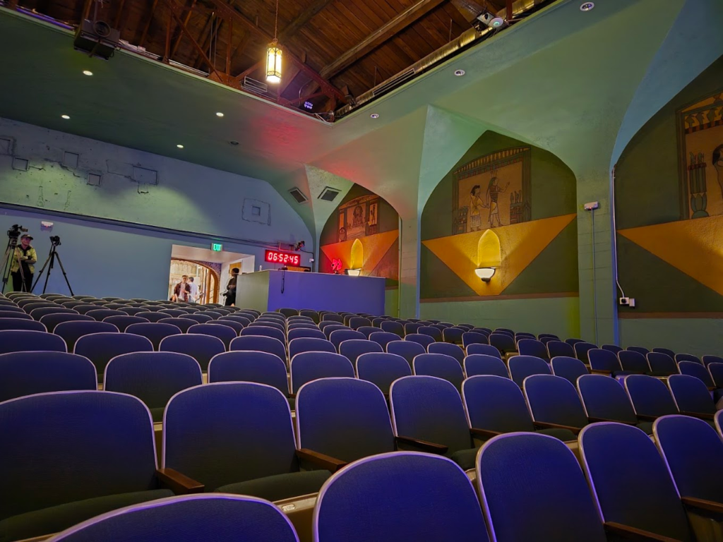 Rows of purple upholstered seats inside a historic movie theater with arched walls, decorative murals and an exposed wooden ceiling.